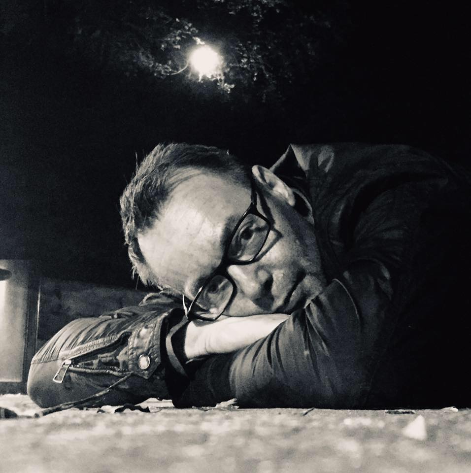 Tom Ole Alvær med briller hviler hodet på armene mot et bord, fotografert om natten med en lyskilde i bakgrunnen. Svart-hvitt.