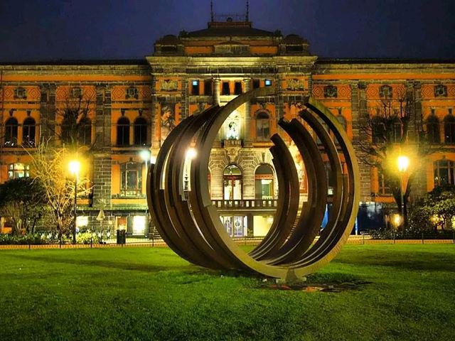 Skulptur av buede metallringer på en plen foran en opplyst murbygning om kvelden.