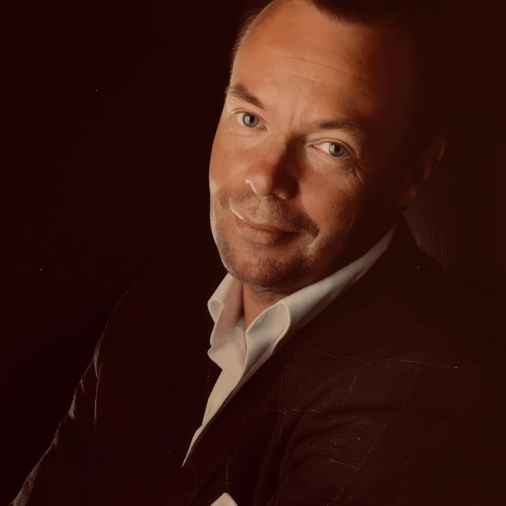 Portrett av Tom Ole Alvær i mørk jakke og hvit skjorte mot en mørk, varm bakgrunn.